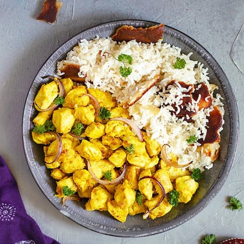 A flat lay of Indian chicken dish in a shallow gray bowl on one side and white rice on the other on a gray background and a purple towel