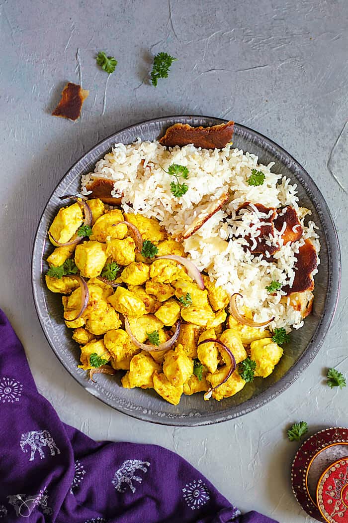 A flat lay of Indian chicken dish in a shallow gray bowl on one side and white rice on the other on a gray background and a purple towel