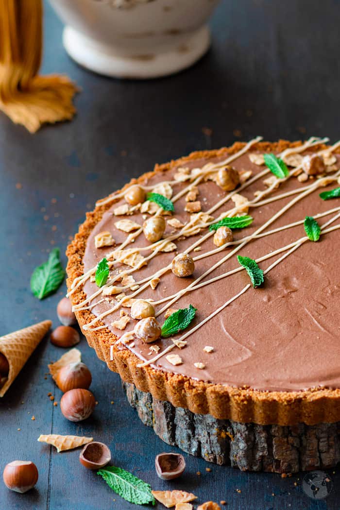 An angle shot of a mousse tart sprinkled with hazelnuts and mint leaves on a wooden plank.