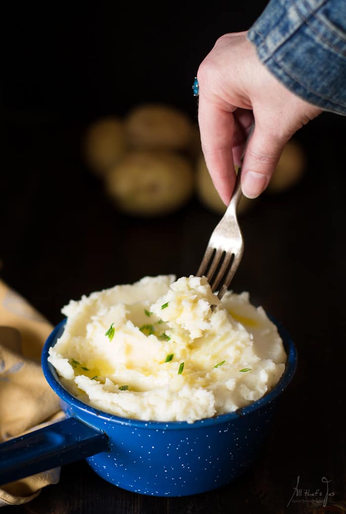 A photo of a blue pot with mashed potatoes being fluffed with a fork. Also in the photo a hand and potatoes in the background.
