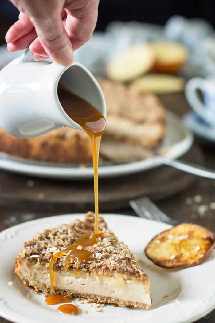 Drizzling caramel over a slice of streusel cheesecake on a white plate with the rest of the dessert in the background.