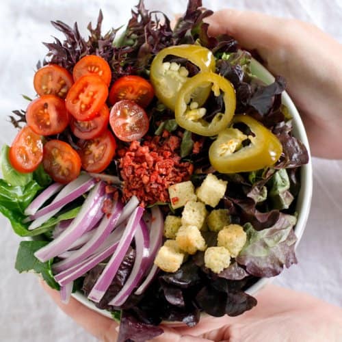 A person holding a bowl with lettuce, tomatoes, onions, peppers, and croutons