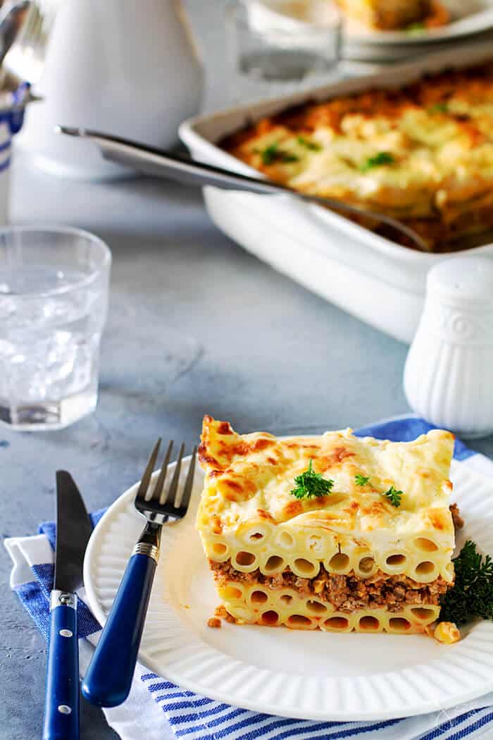 A serving of noodle moussaka on a white plate on blue-gray background and white dishes.