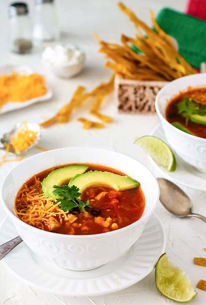 A white bowl on a white background filled with chicken soup and topped with avocado slices.