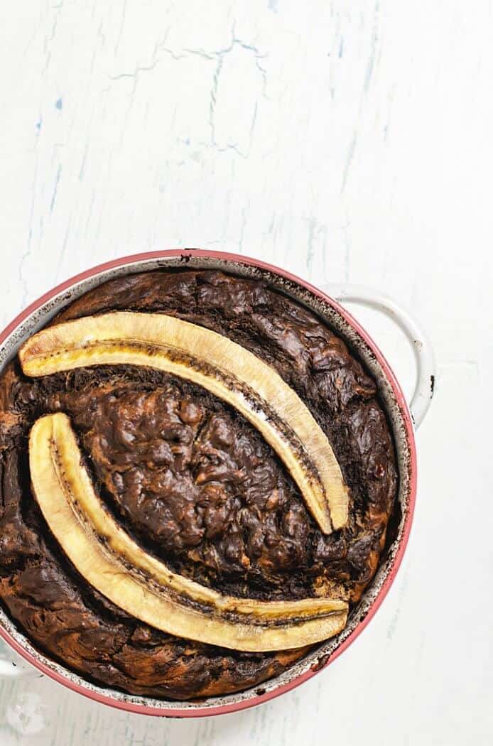 A round pan with chocolate banana bread topped with two banana halves on a white counter.