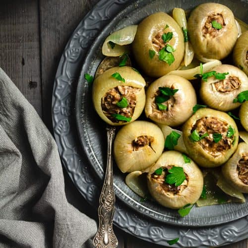 An overhead shot of stuffed onions on a gray plate with a spoon and gray towel on the side. Also in the photo - lemon wedges.