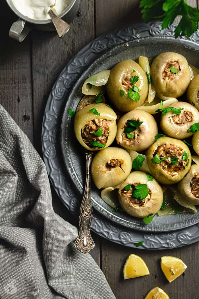 An overhead shot of stuffed onions on a gray plate with a spoon and gray towel on the side. Also in the photo - lemon wedges.