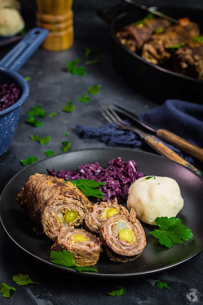 black plate on a dark blue background with rouladen, red cabbage, and dumplings.