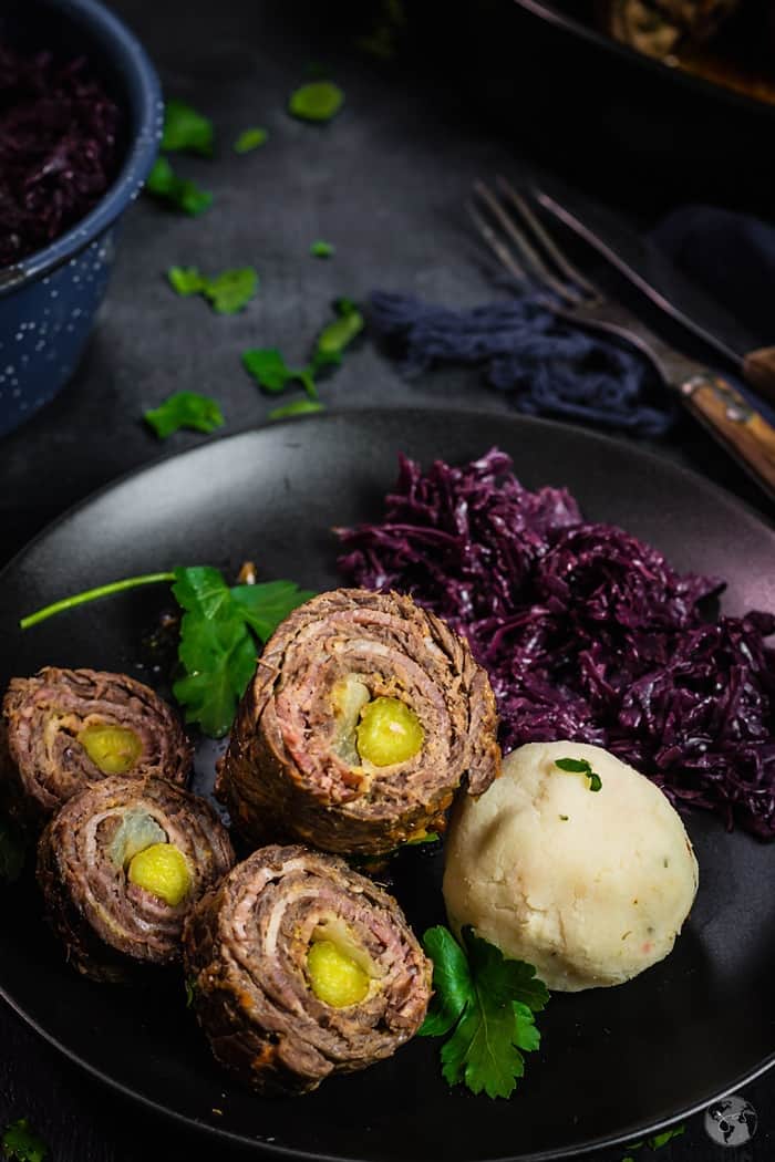 A shot of a sliced German beef roll ups served on a black plate with red cabbage and dumpling, on a dark background