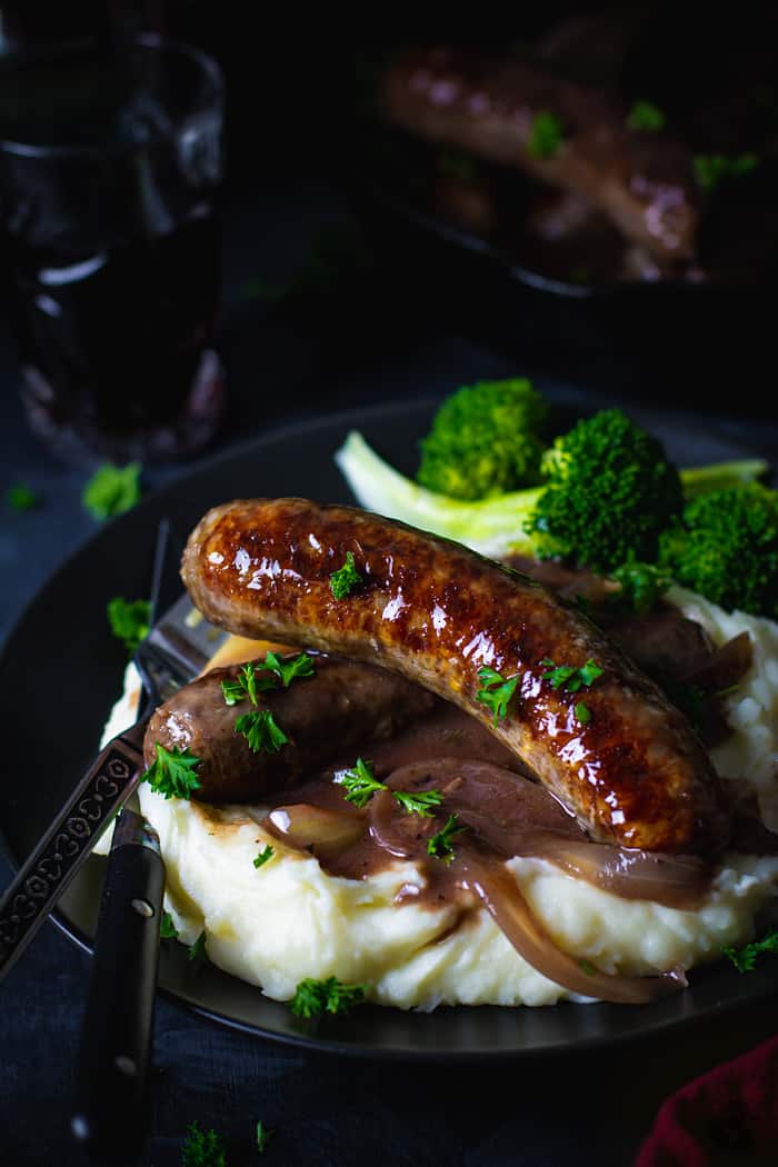Two brats in gravy over mashed potatoes and broccoli on a black plate