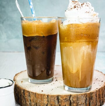 Two glasses with Greek frappe on a round slab of wood and light blue background.