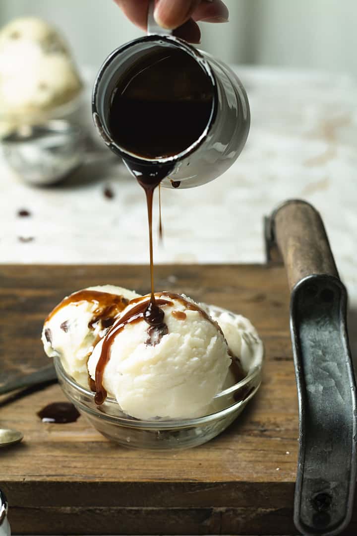 A bowl with three ice cream scoops on a wooden tray and rum sauce being poured over.