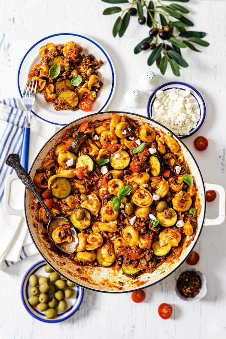 An overhead shot of Greek beef and cheese tortellini dish in a cast iron enameled skillet on a white background and servings of feta and olives.