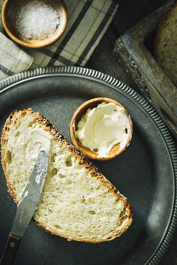 A flat lay of one slice of bread on a gray round metal plate with a knife and two bowls with butter and salt.