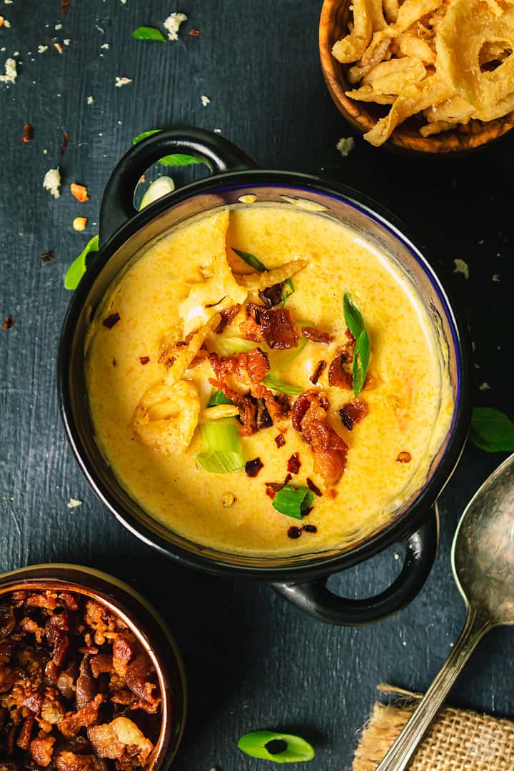 An overhead shot of a cheese dish in a black mini pot with extra bacon and fried onions in separate bowls.