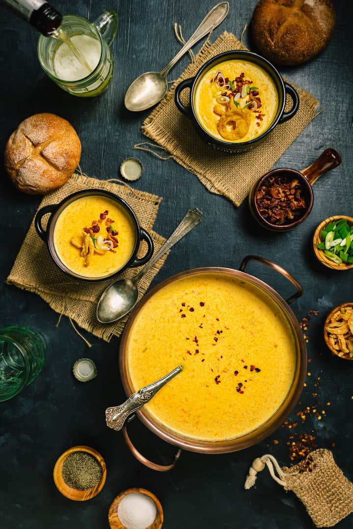 A flat lay of a pot with beer cheese soup with two servings, beer glasses, and small bowls with garnishes on a dark background.