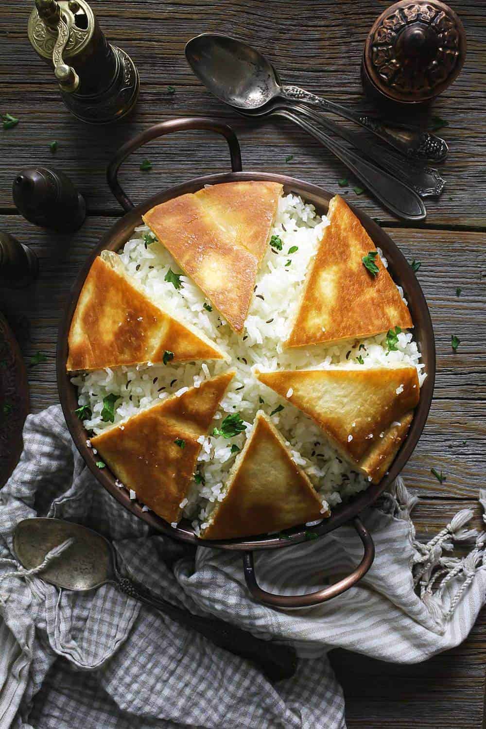 A copper bowl filled with rice and covered with crispy naan bread pieces sitting on a wooden table.