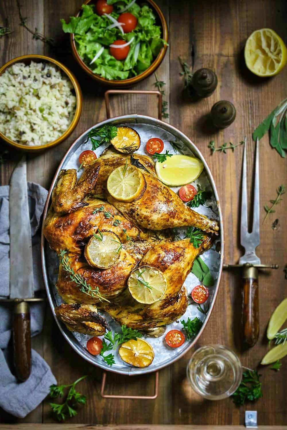 An oval dish with roasted whole chicken with lemon slices and fresh herbs sitting on a wooden table with cutlery and side dishes.