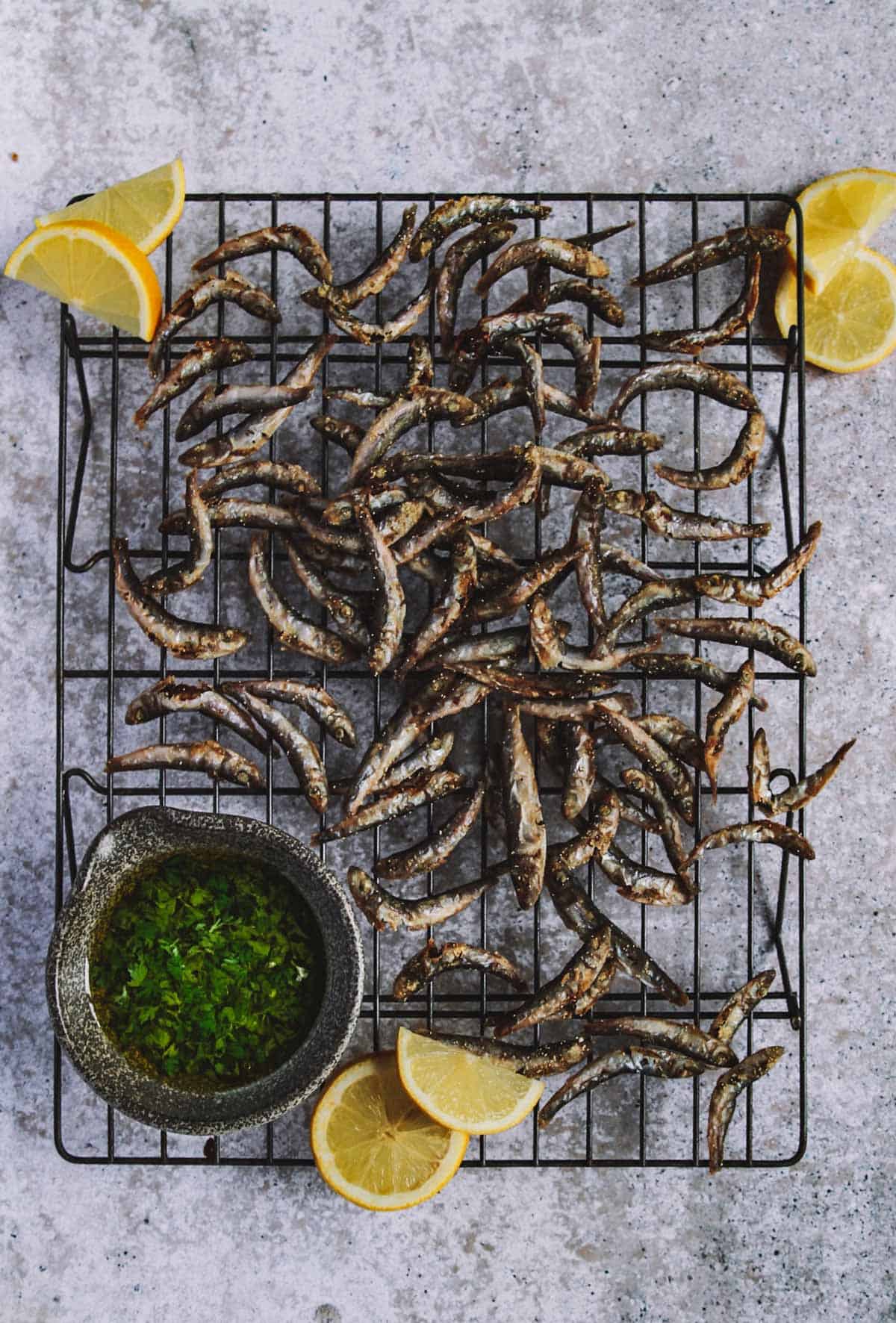 A cooling rack on a table with small fried fish, lemon slices and a bowl with sauce.