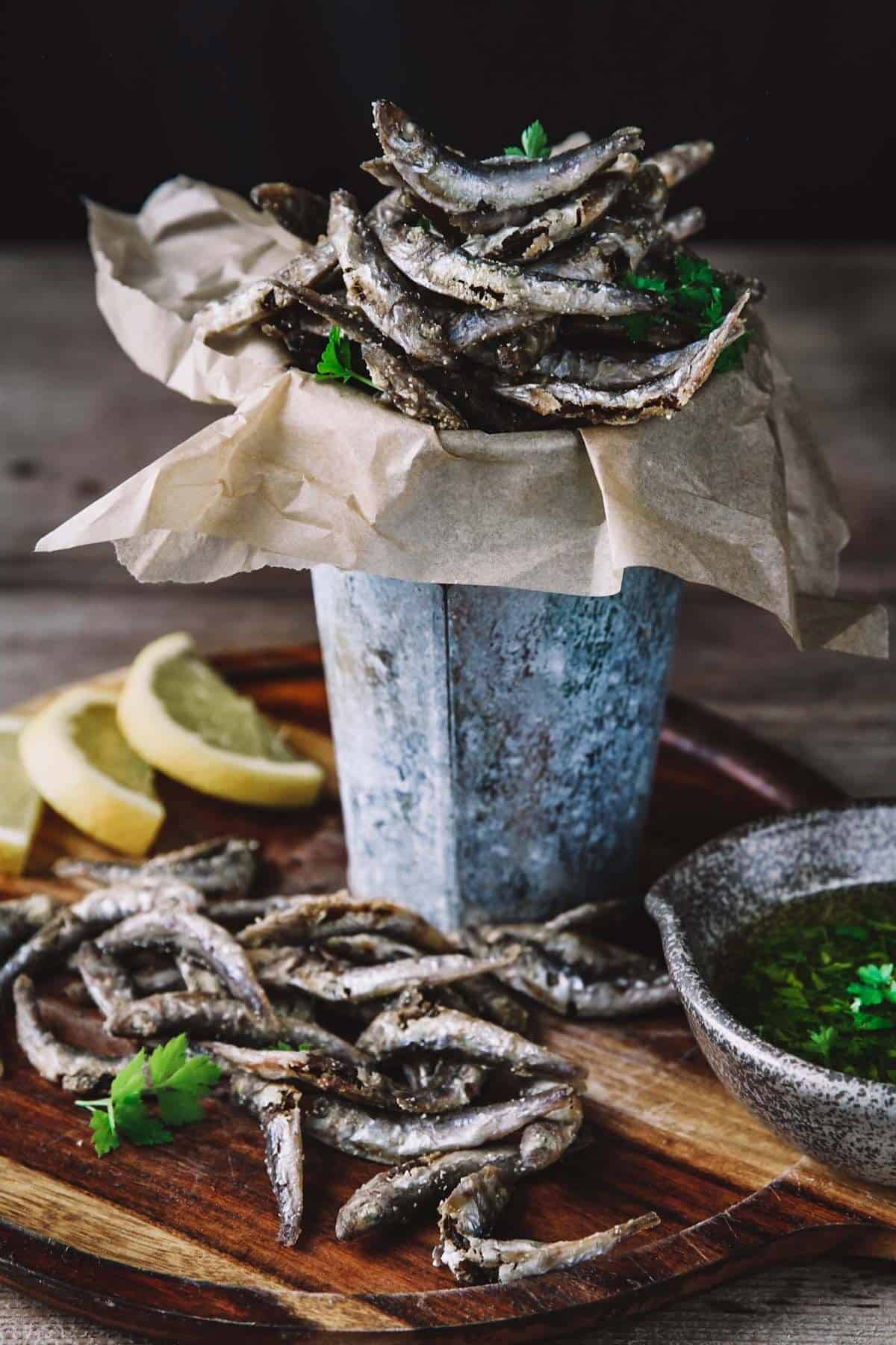 A metal cup filled with fried fish sitting on a wooden board surrounded with more fish, lemon, and sauce.