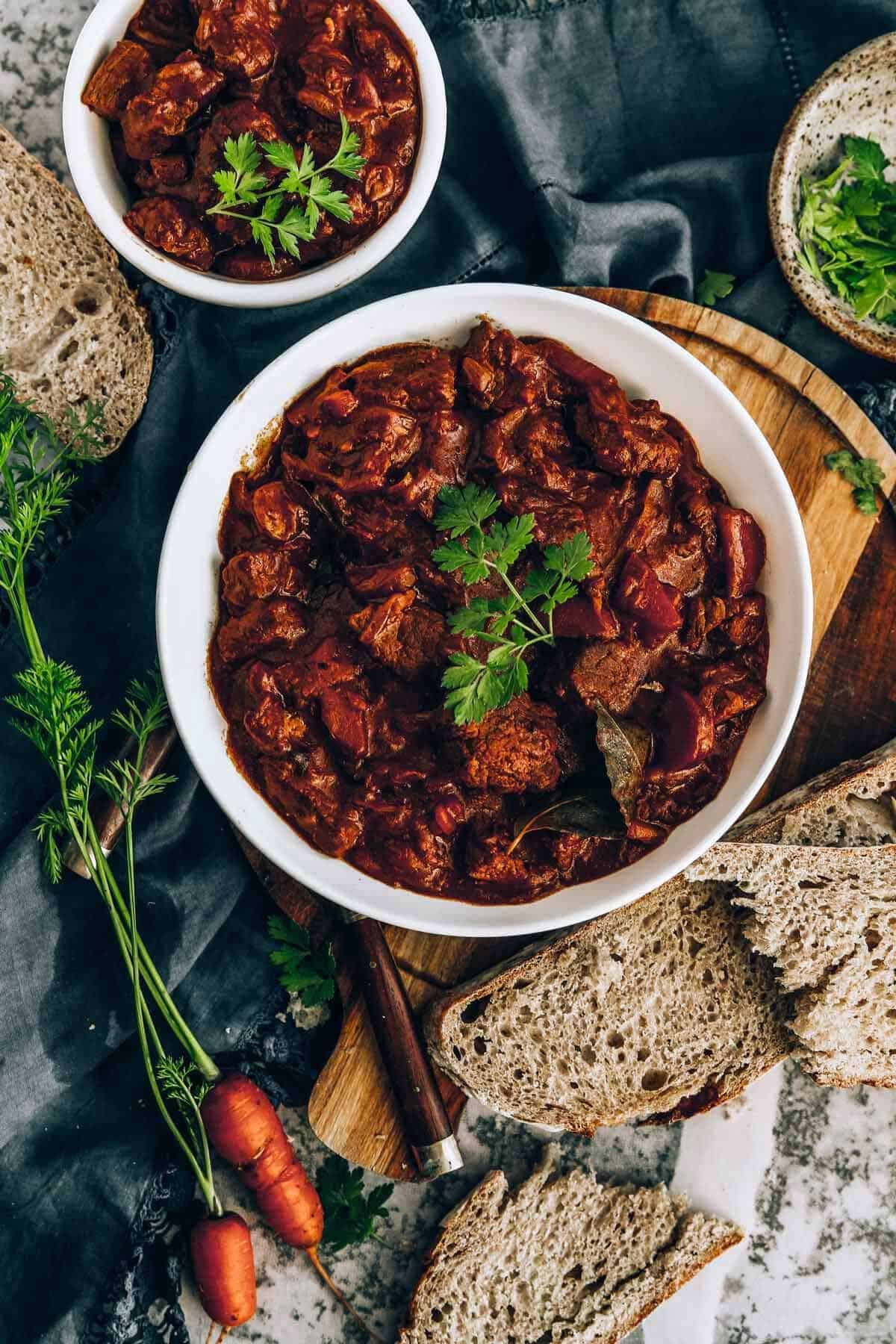 Two bowls with beef stew sitting on a rustic table with bread and carrots