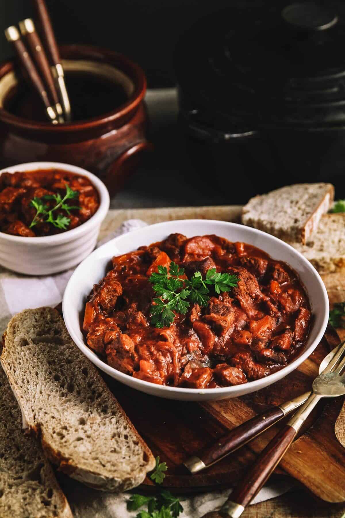 A lage and a small plaate of beef stew on a wooden table with bread and utensils