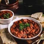 A lage and a small plaate of beef stew on a wooden table with bread and utensils