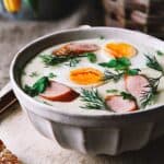 A bowl of white Polish creamy soup with boiled egg halves and kielbasa sausage.