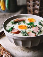 A bowl of white Polish creamy soup with boiled egg halves and kielbasa sausage.