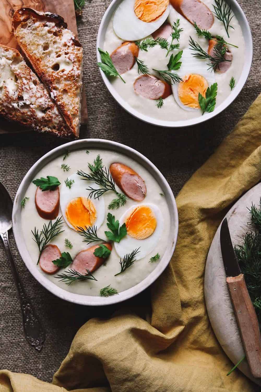 Two bowls of Polish bialy barszcz topped with hard-boiled eggs halves, sliced sausage, and fresh dill. 