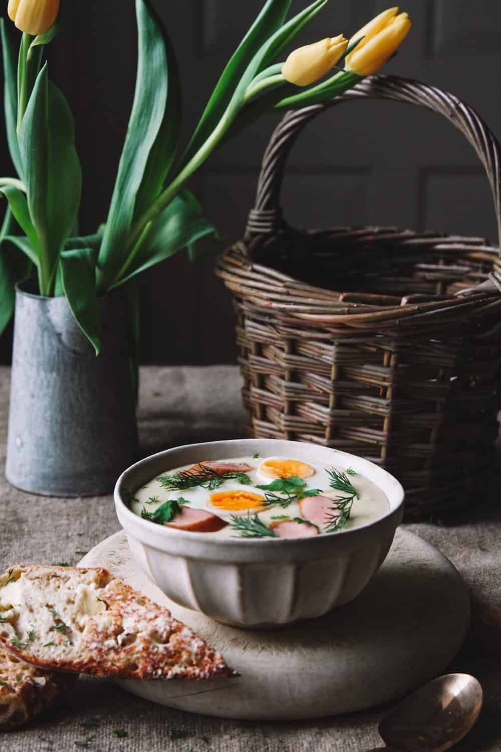 A bowl with Easter breakfast soup traditional to Poland on a table with a basket and tulips in a vase.