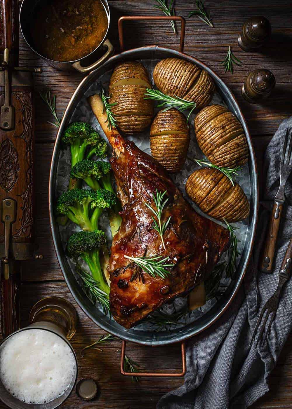 Slow roasted leg of lamb with beer on a metal roasting pan with potatoes and broccoli on a wooden table.