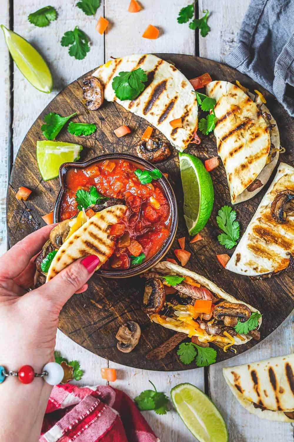 A woman dipping steak quesadilla into a bowl with salsa with more quesadillas spread on a wooden plank.