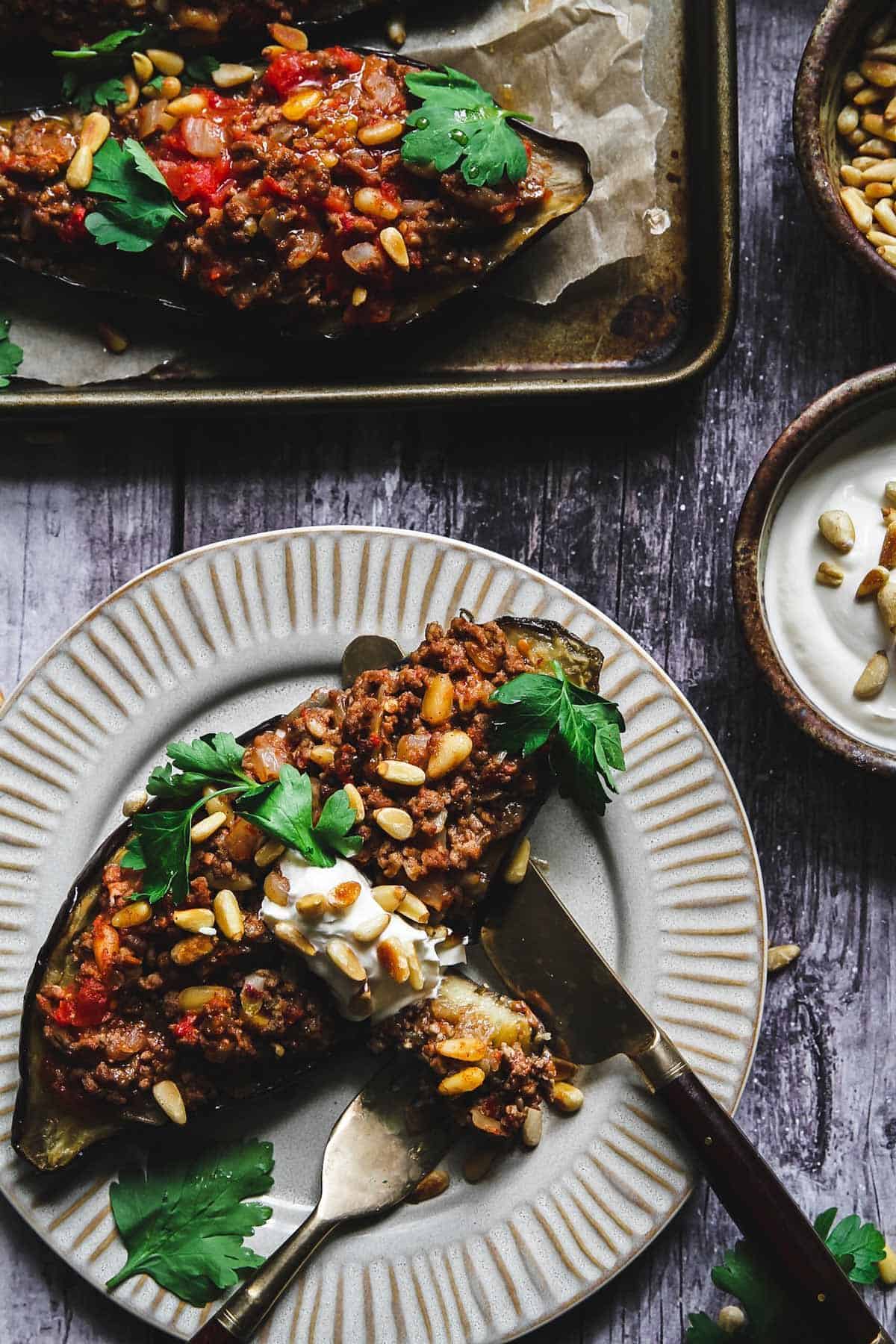 Roasted eggplant half stuffed with ground lamb and pine nuts topped with sour cream and parsley on a white plate.