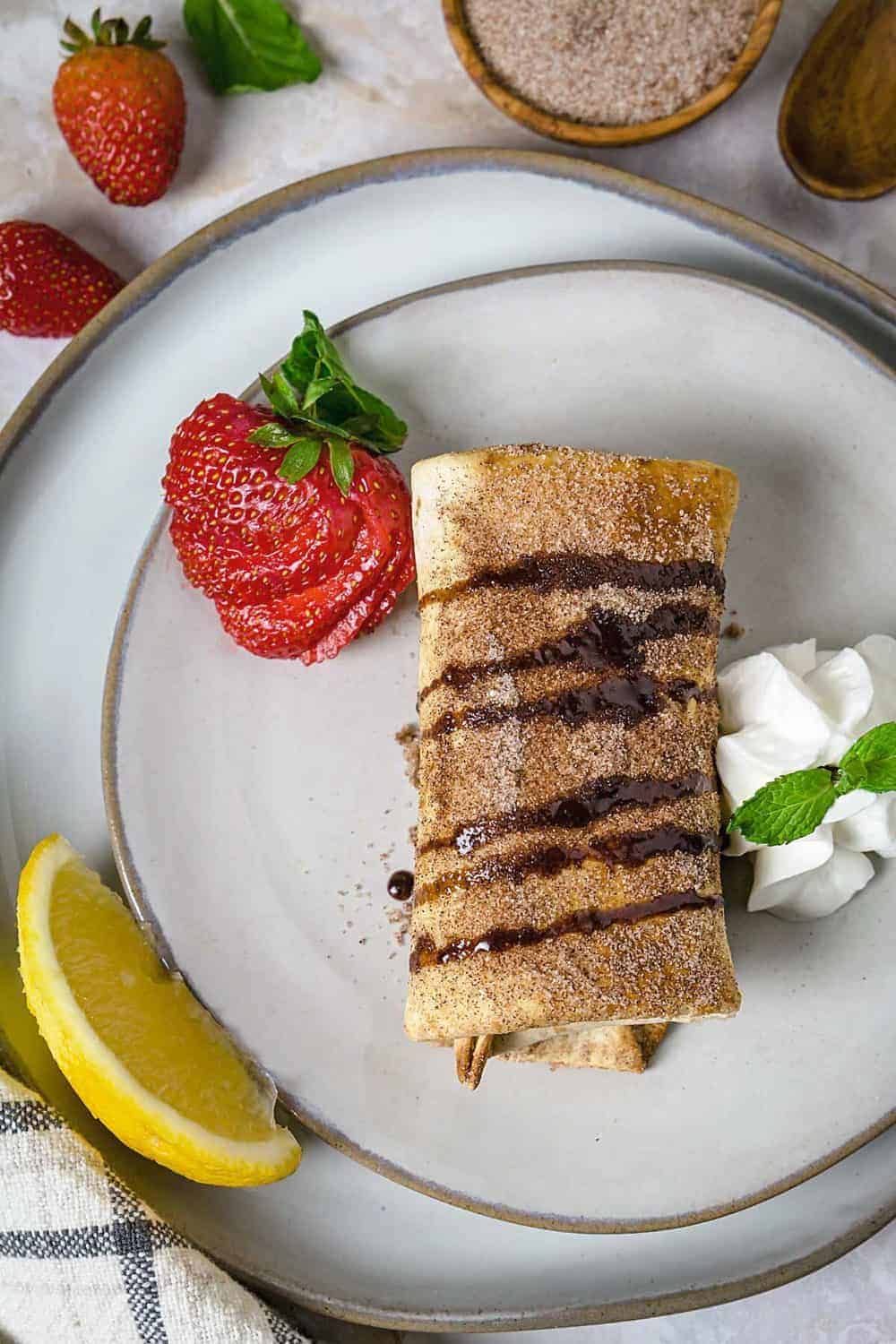 Chimichanga on a plate with chocolate drizzle, lemon, whipped cream, and strawberry.