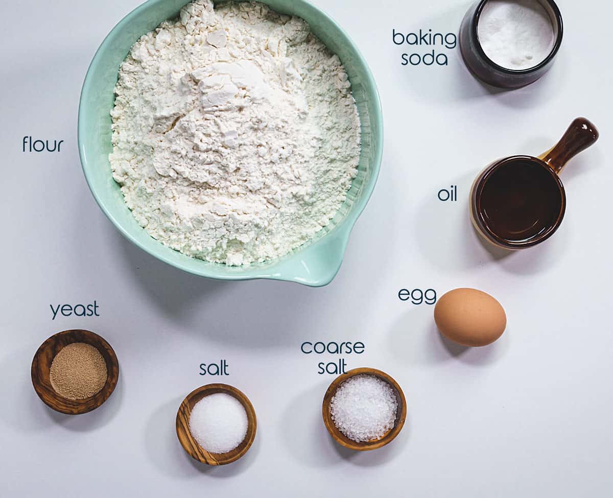 Ingredients for pretzel rolls on a table with flour, yeast, salt, egg, and baking soda.