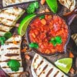 Small grilled tortillas with steak, mushrooms, and lime wedges placed around a bowl with salsa.