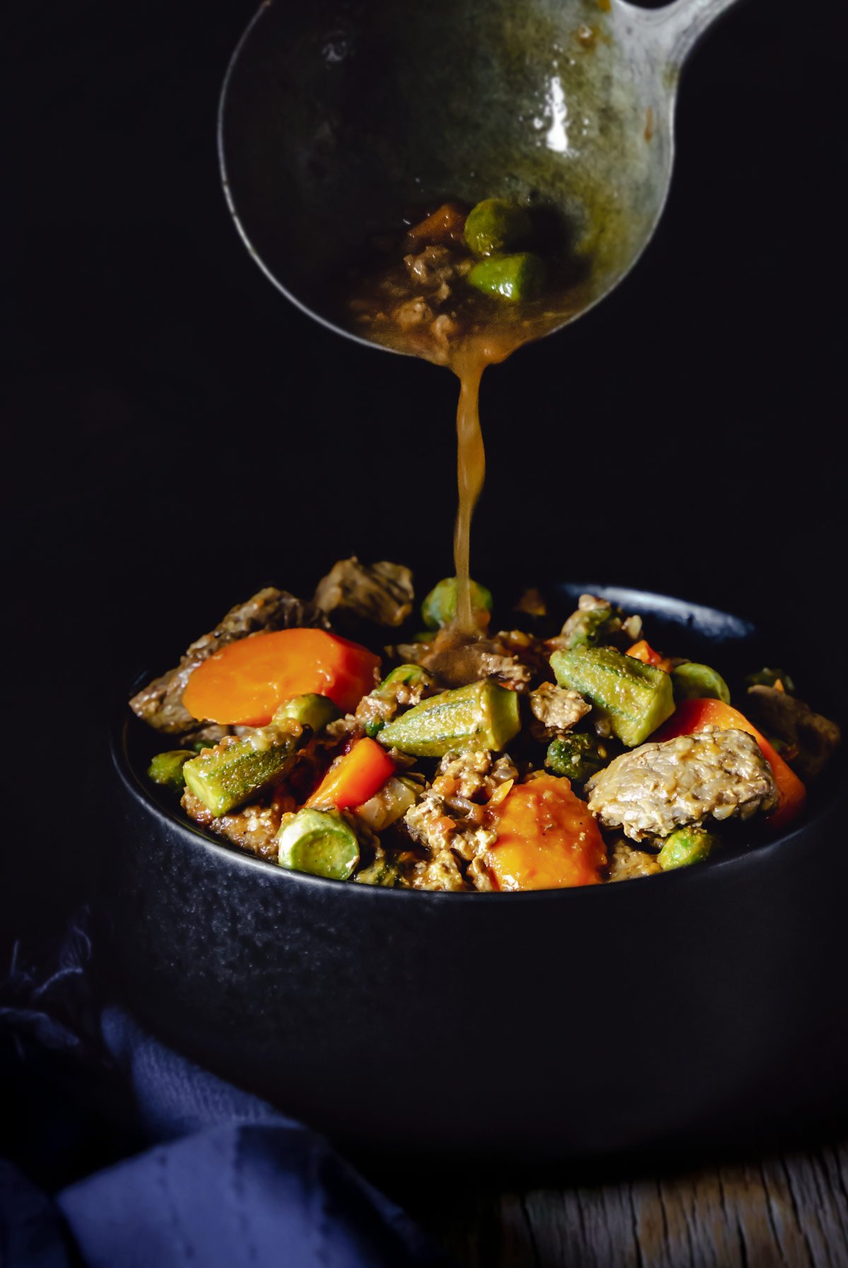 A ladle pouring the okra stew over a black bowl with meat and vegetables.