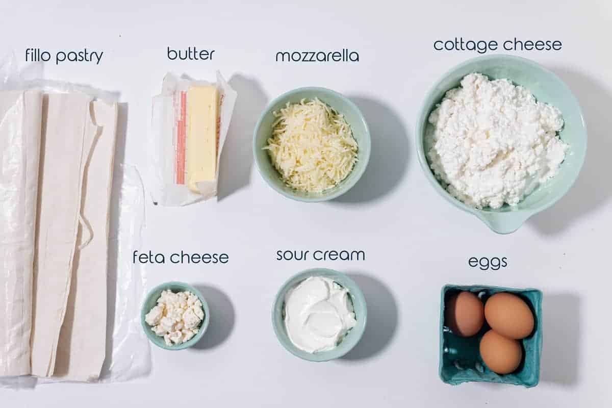 Ingredients for cheese pie with fillo dough, eggs, and three types of cheese on the counter.