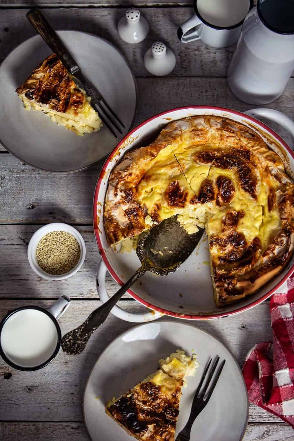 Round baking dish with fillo egg pie and two slices on plates sitting on a wooden white table.