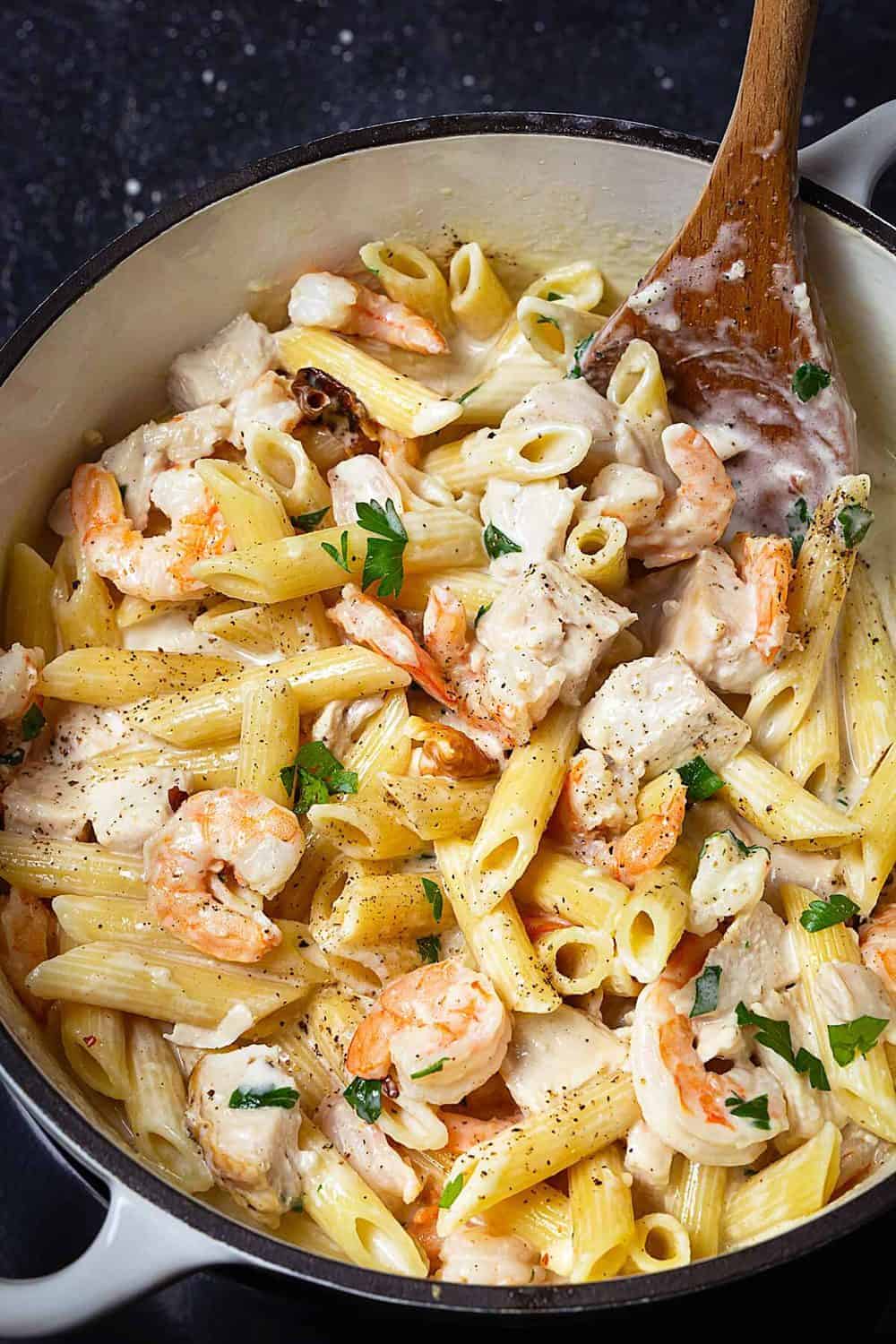 A Dutch oven with creamy alfredo pasta, chicken pieces, and shrimp with a wooden spoon in it.