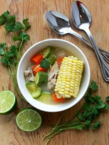 A bowl of Mexican chicken soup with corn on the cob, veggies, and lime for serving.