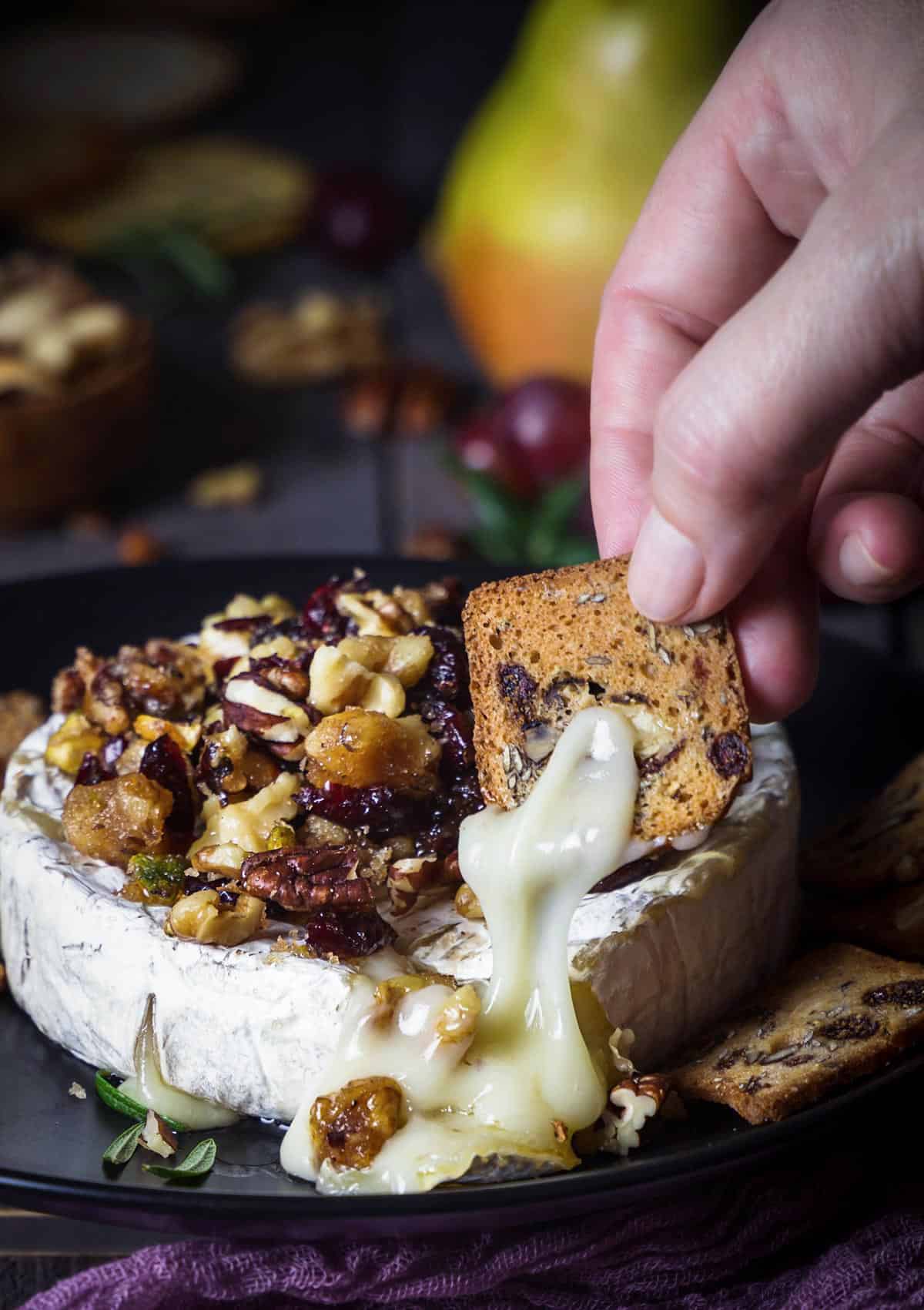 A person dipping a cracker into the melted brie cheese with walnuts, pistachios, and pecans.