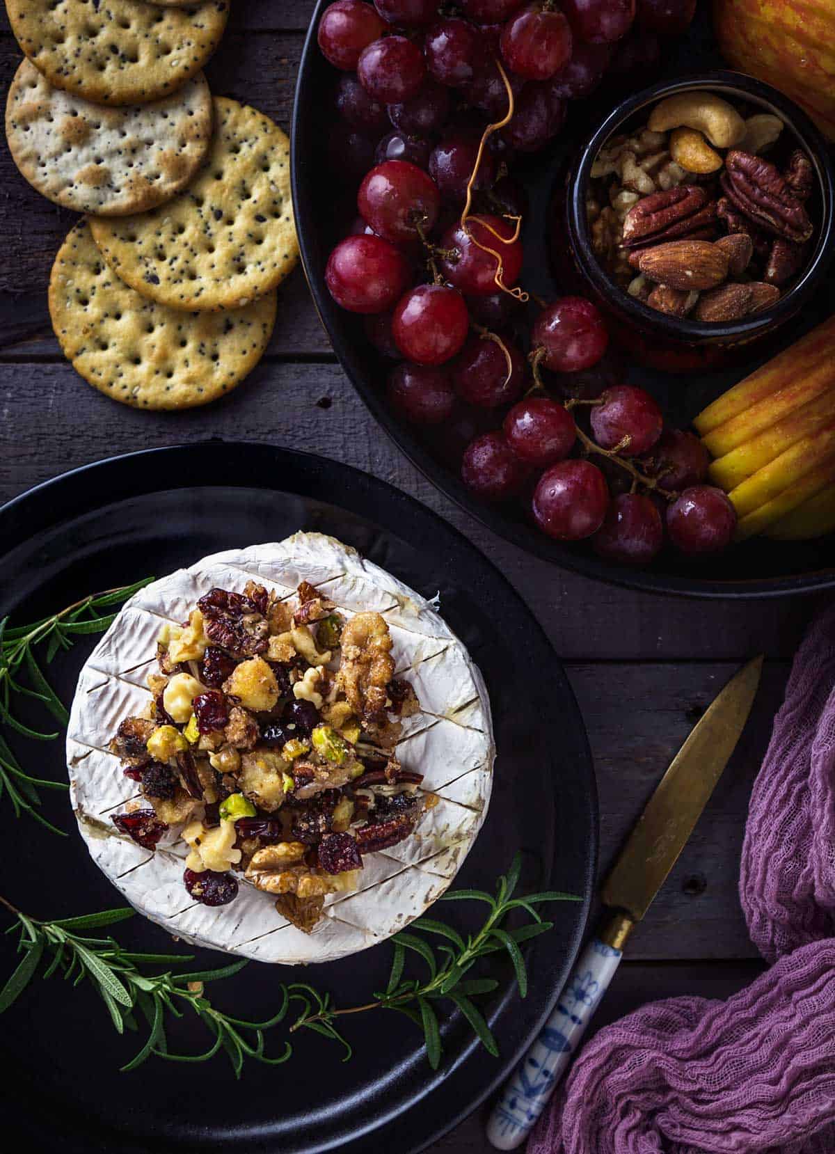 A wheel of baked brie with mixed nuts and cranberry topping on a table with fruit and crackers.