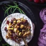A person cutting into a brie with cranberry nut garnish.