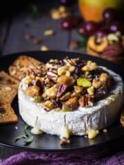 A wheel of camembert or brie on a black plate with toppings oozing o the sides.