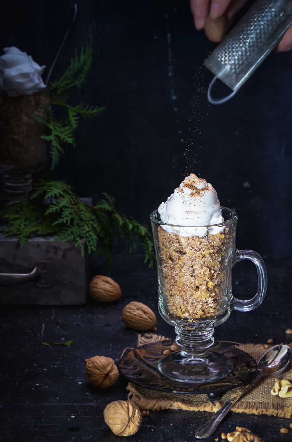 A person grating whole nutmeg over the whipped cream in a glass with wheat pudding.