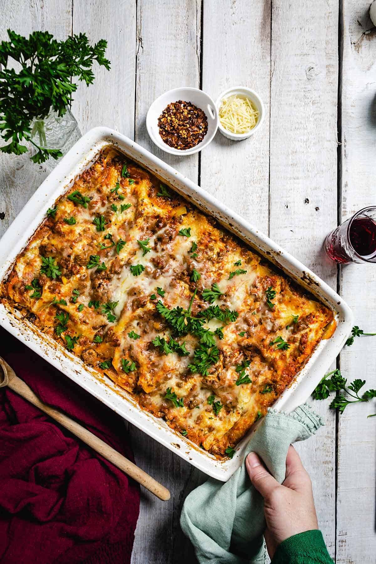 A person putting a white dish with lasagna on a rustic white table.