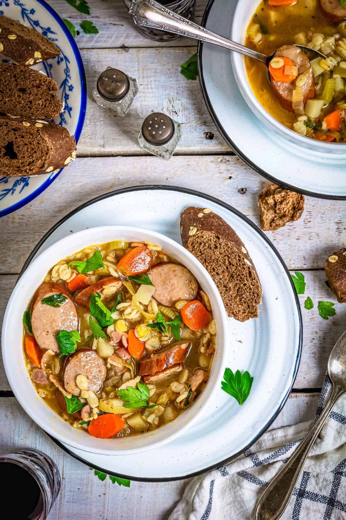 Servings of barley sausage and vegetable soup with bread on a table.