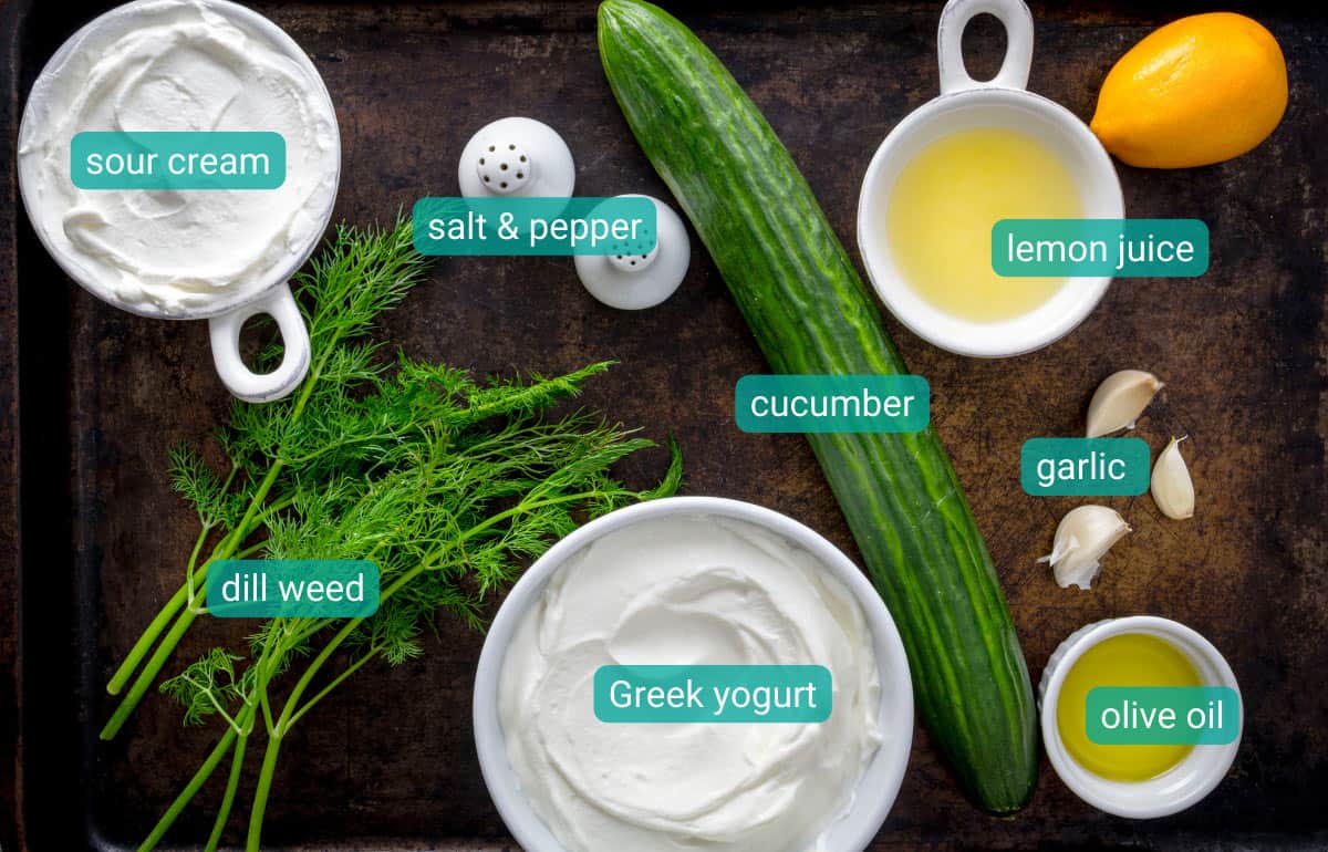 Ingredients for tzatziki dip with cucumber, yogurt, sour cream, lemon, and dill on the counter.